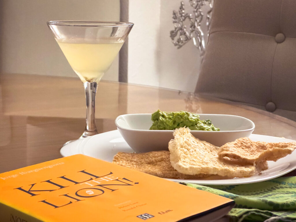 A hardcover book, Kill the Lion by Jorge Ibargüengoitia, sits on a table beside a plate of guacamole and chicharones and a margarita.