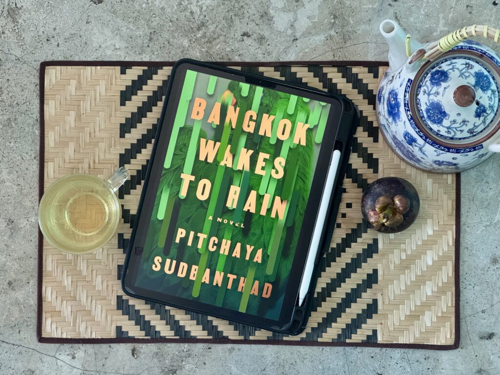Flat lay photo of the eBook cover of Bangkok Waits for Rain displayed on an iPad with a pot and cup of tea and a mangosteen fruit.