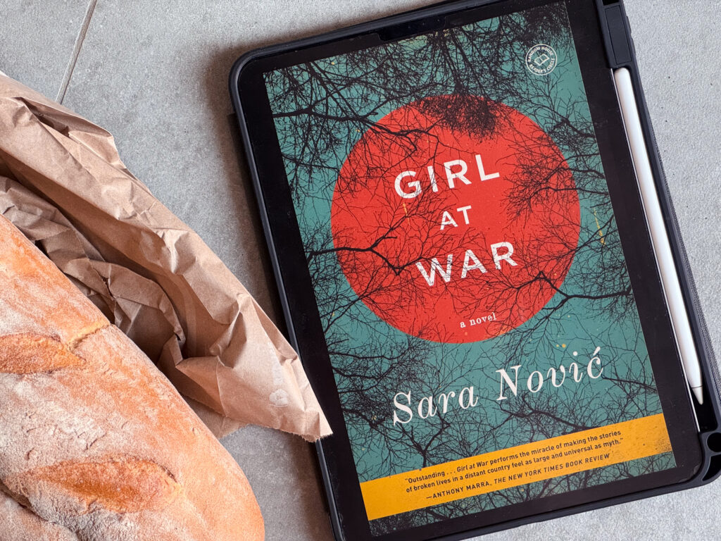 A loaf of bread lying on a table next to an iPad displaying the cover of Girl at War by Sara Novic